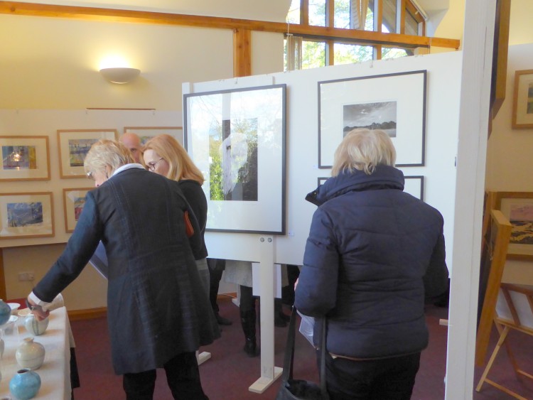 A group viewing Rob Wheeler's ceramics, with Richard Hayward's photographs visible to the right.